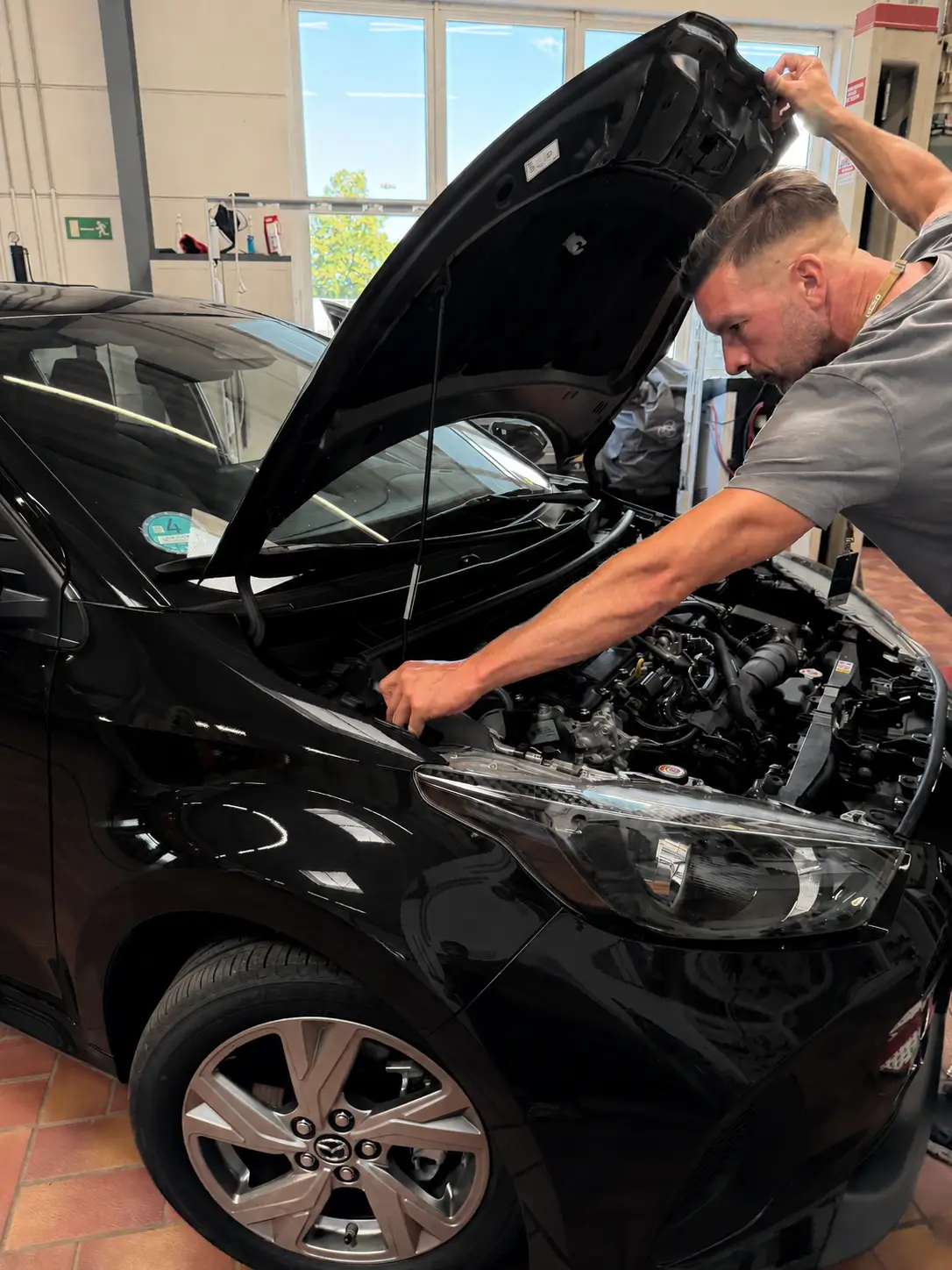 Ein Mechaniker öffnet die Motorhaube eines schwarzen Mazda und greift in den Motorraum.
