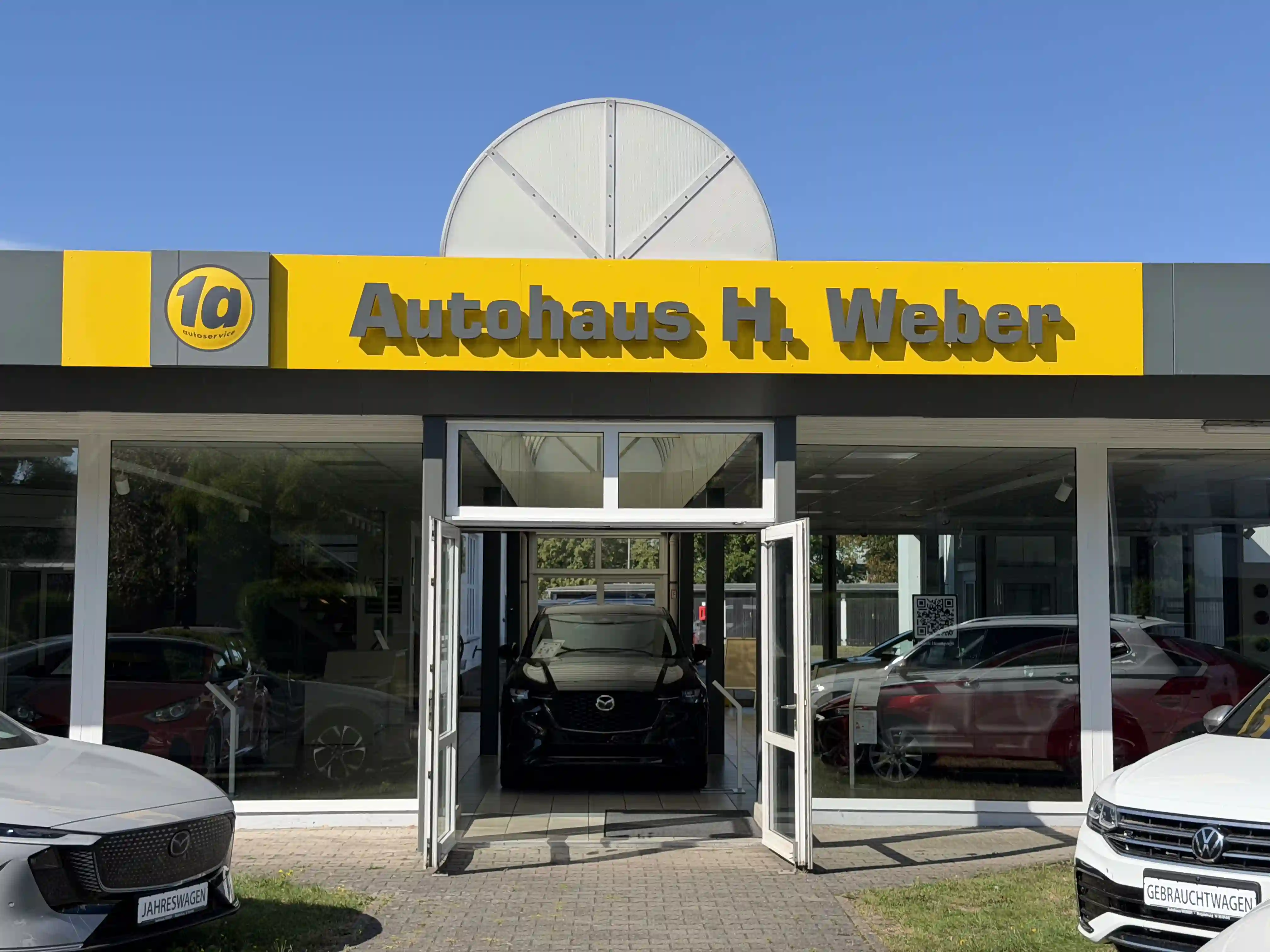 Autohaus H. Weber von der Straße aus: gelber Schriftzug über der Glasfront, offene Eingangstür, davor zwei Fahrzeuge.