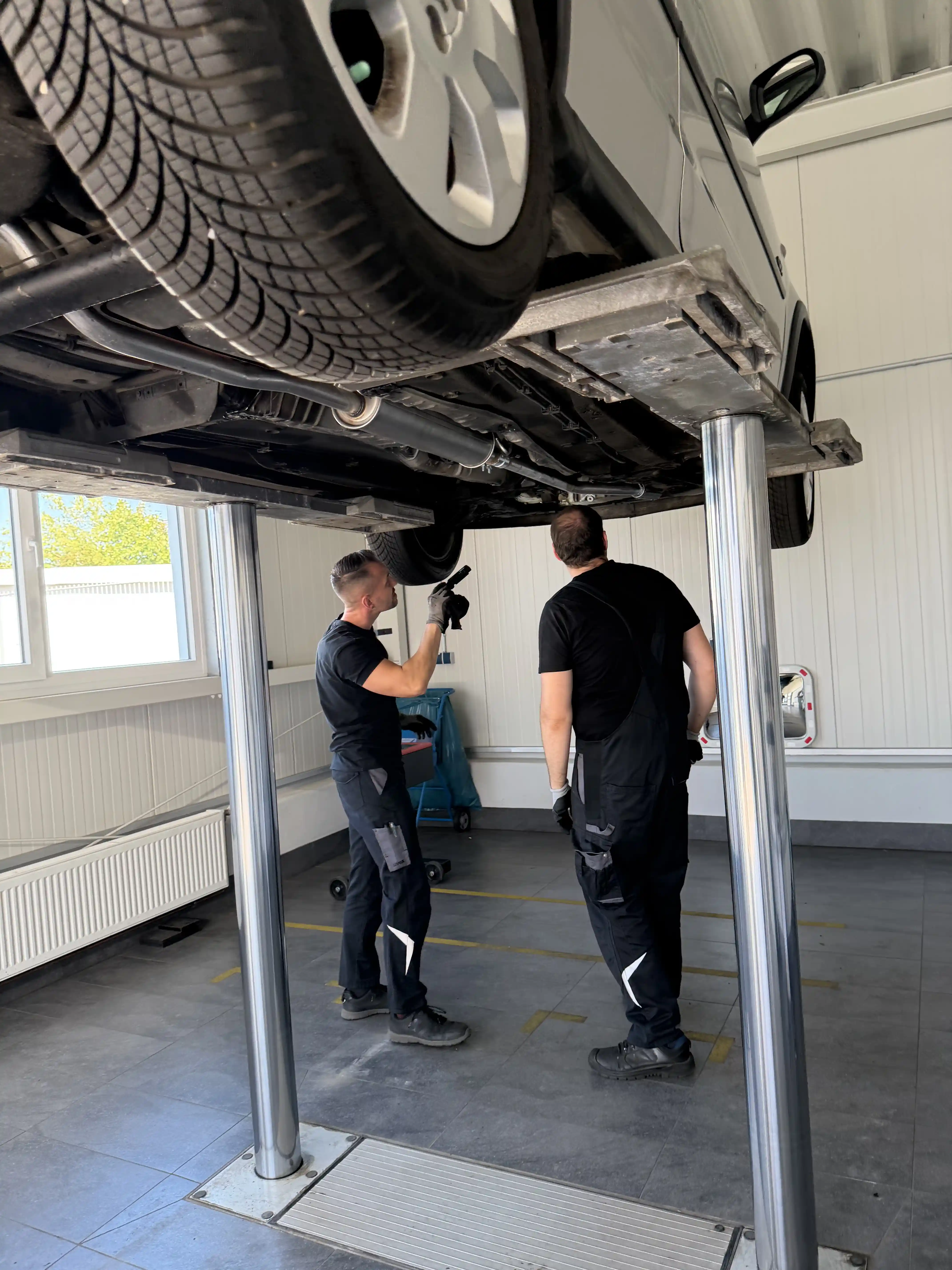 Fahrzeugdurchsicht in Magdeburg: zwei Mechaniker stehen unter einem angehobenen Auto, einer leuchtet mit der Taschenlampe an die Hinterachse.